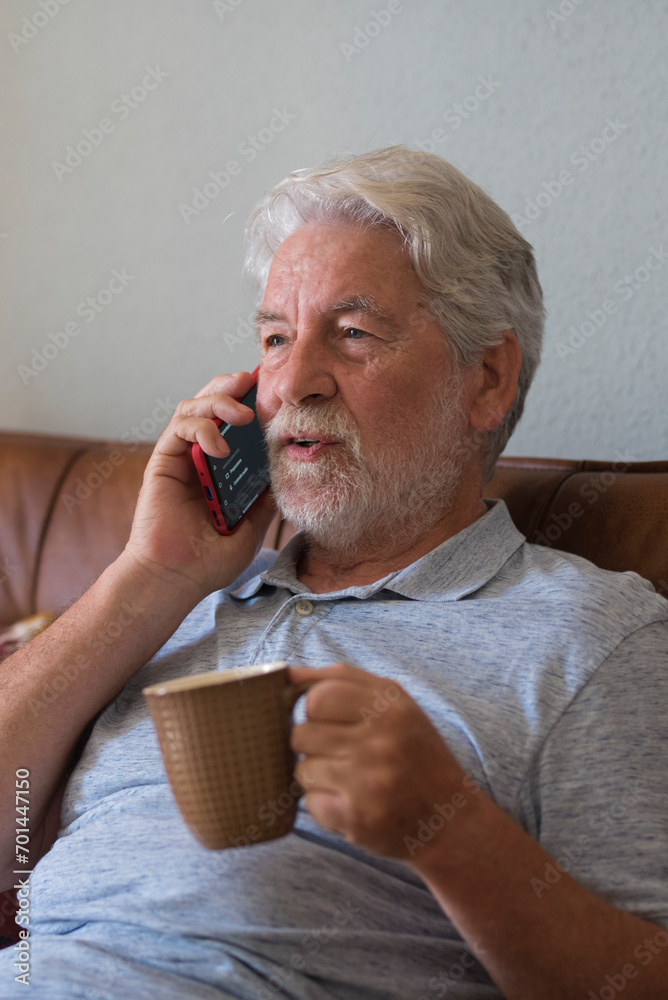 Old senior man at home speaking with modern phone and drinking coffee or tea sitting on the sofa and enjoying technology connection - one mature male use cellular at home relaxing and having fun Old senior man at home speaking with modern phone and drinking coffee or tea sitting on the sofa and enjoying technology connection - one mature male use cellular at home relaxing and having fun
