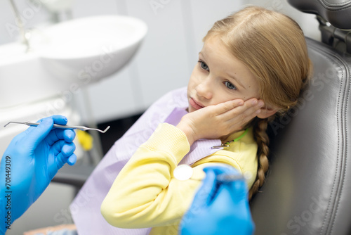 Sad child girl sitting in dentist's chair and holding hand to his cheek from toothache and looking at doctor. Kid dental problems photo