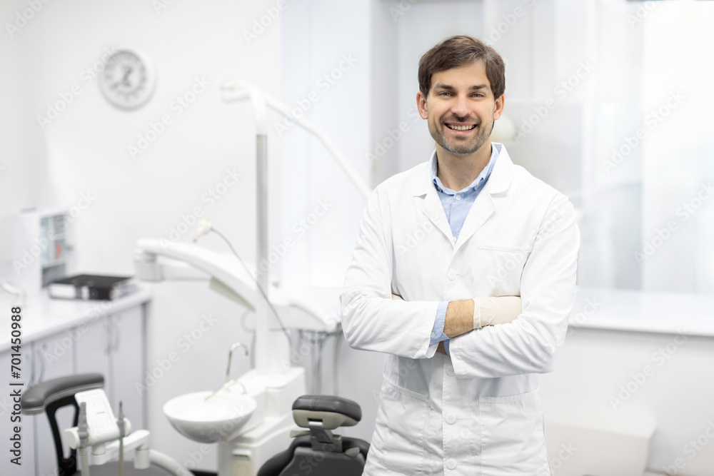 Dental Center. Portrait of male dentist doctor posing at workplace, stomatologist standing with folded arms in modern clinic interior