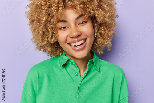 Portrait of good looking woman with blonde curly hair smiles toothily expresses postive emotions wears earrings and green jumper isolated over purple background. People and happiness concept photo