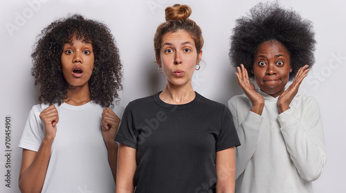 Photo of shocked European woman with hair bun wears casual black t shirt poses between two African women with curly hair feel impressed isolated over white background. Collage shot. Human reactions