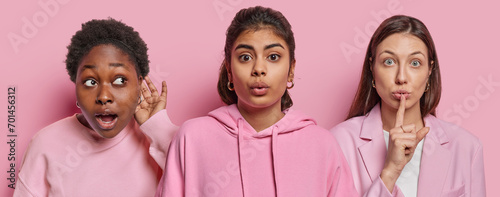 Photos Horizontal shot of three women stand next to each other one tries to overhear something other makes silence gesture and third reacts to something surprising isolated over pink studio backgrund