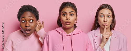 Horizontal shot of three women stand next to each other one tries to overhear something other makes silence gesture and third reacts to something surprising isolated over pink studio backgrund. photo