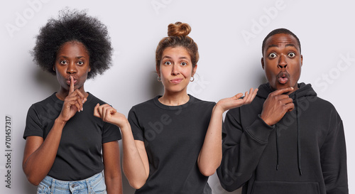Horizontal shot of hesitant European woman spreads palms with clueless expression dark skinned woman makes silence gesture African man reacts stupefied dressed in black clothing isolated on white wall