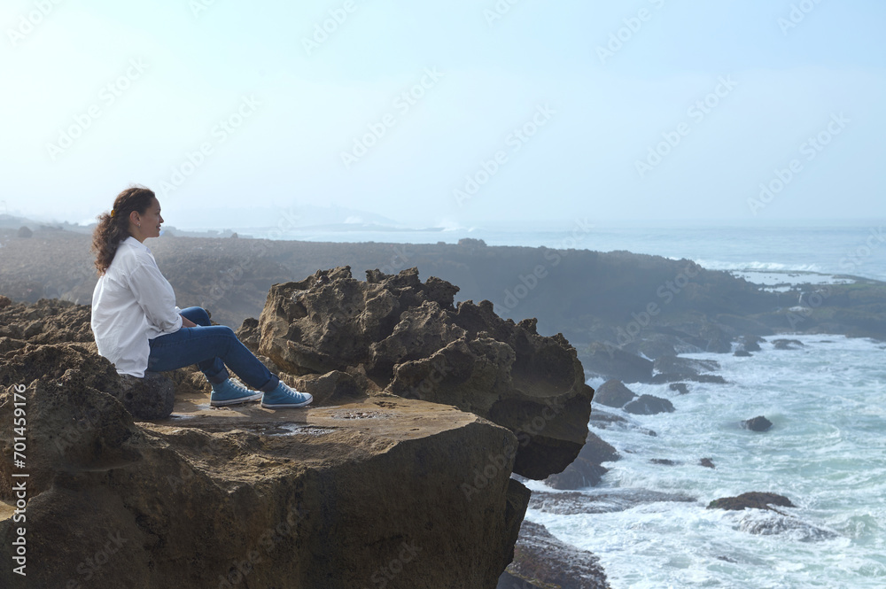Relaxed multi ethnic woman sitting alone on a cliff and admires the mesmerizing view of the Atlantic Ocean.