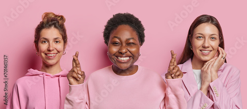 Studio shot of three women stand next to each other smile happily keep fingers crossed believe in good luck express positive emotions isolated over pink background. People and body language concept