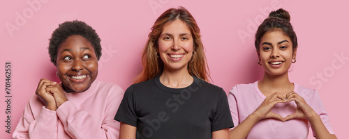 Three positive women smile gladfully make heart gesture express love focused aside with dreamy expression dressed in casual clothing isolated over pink background. Collage image of female friends photo