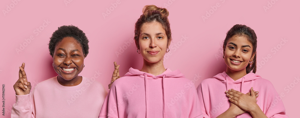 Horizontal shot of three women stand closely to each other keep fingers crossed believe in good luck press hands to heart feel thankful dressed in casual clothing isolated over pink background. Horizontal shot of three women stand closely to each other keep fingers crossed believe in good luck press hands to heart feel thankful dressed in casual clothing isolated over pink background.