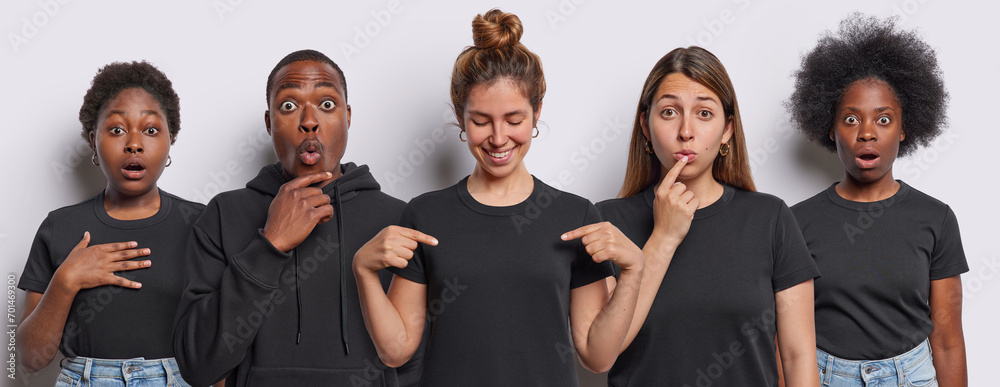 Image of shocked women and one African American man stare at camera with widely opened eyes one cheerful woman points at mock up space of casual black t shirt isolated over white background.