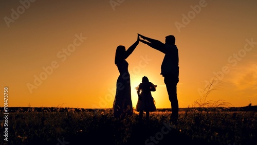 Silhouette of daughter standing between parents forming figure with hands similar to house for family. Silhouette of little daughter standing under father and mother arms forming dream house at sunset