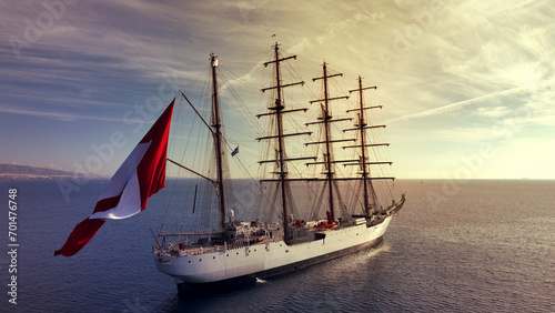 Aerial drone photo of beautiful three mast barque or barc type classic sailing wooden boat with huge waving flag anchored in Mediterranean deep blue Aegean sea at sunset with golden colours