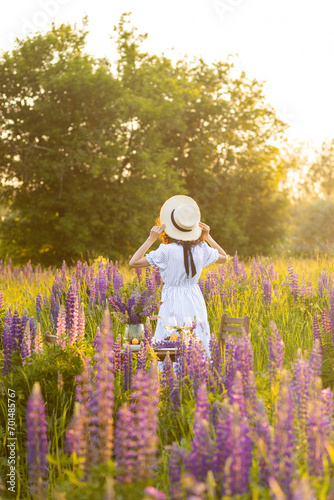 Golden hour, sunset, summer. Beautiful happy young woman on meadow arranging table for outdoor event, gathering wildflowers. Wedding or romantic date decoration in the field with purple lupins