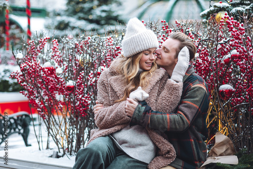 Quadro su tela Beautiful young loving couple embracing, kissing, enjoying winter vacation, cold snowy day on a street city Christmas market outdoors