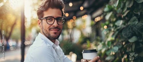 Professional modern and confident young businessman with glasses and white shirt holding a eco friendly paper cup of coffee in urban park of business district. Creative Banner. Copyspace image photo