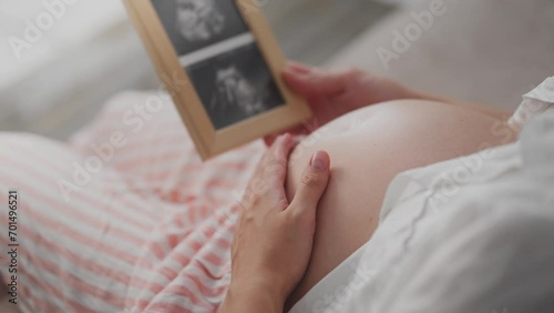 A pregnant girl in loungewear looks at a photo of her unborn child's bridles. White woman holds in her hands a frame with a photo of the reins caring for the future of mother and baby, child photo