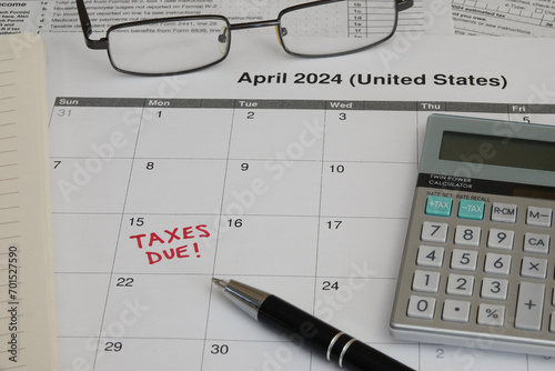A 2024 calendar noting the April 15 USA Internal Revenue Service IRS income filing deadline for year 2023 taxes is shown up close, with a calculator, ink pen, notebook, and glasses in the frame.