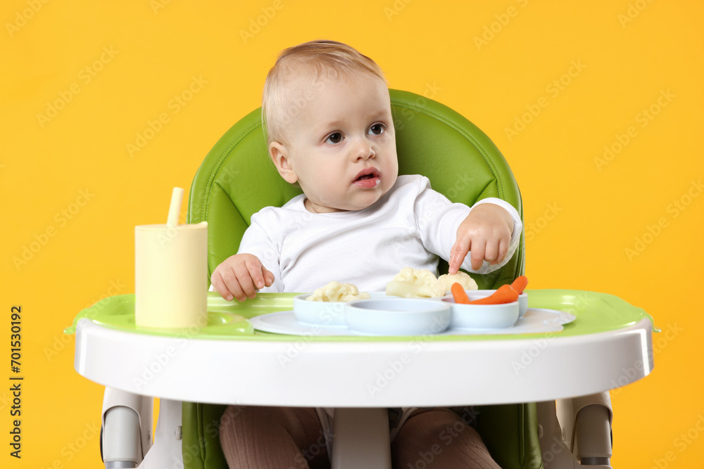 Cute little baby with healthy food in high chair on orange background Cute little baby with healthy food in high chair on orange background