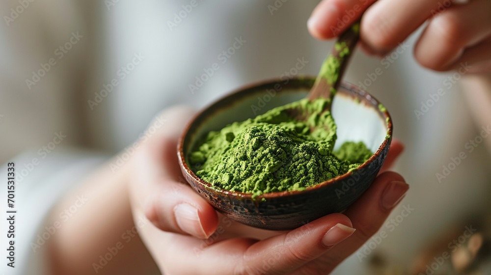 Woman in white prepare Japanese green Matcha tea by scooping green tea powder out of a traditional ceramic bowl on a blurred green white background with copy space.