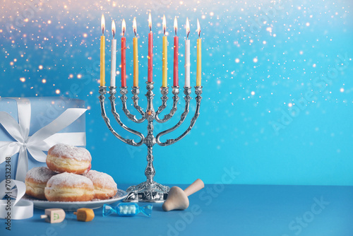 Hanukkah celebration. Menorah with burning candles, dreidels, donuts and gift box on light blue table, space for text