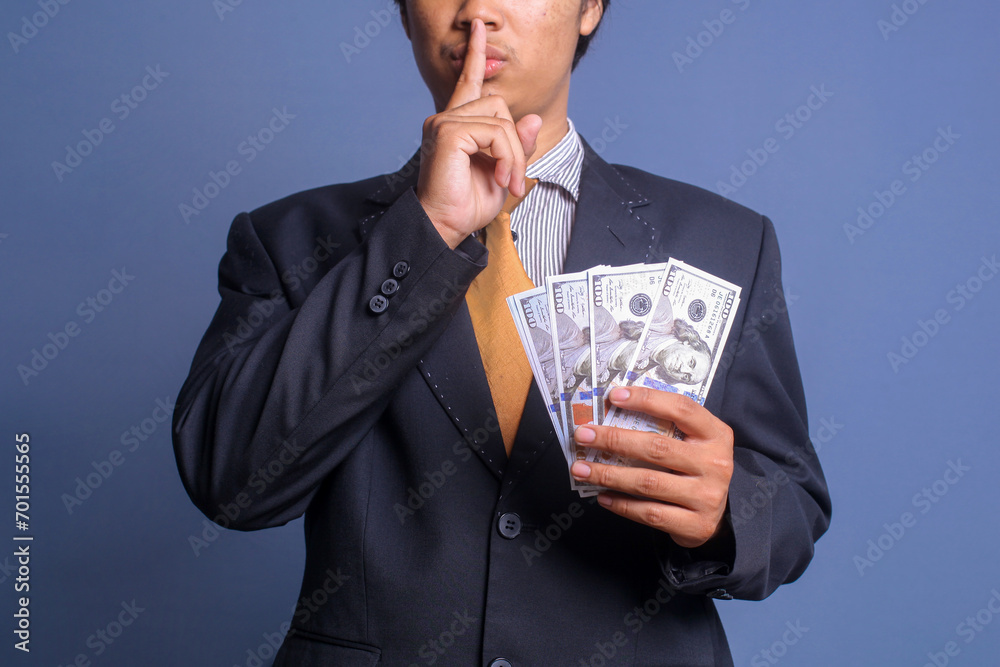 Man in suit put finger on lips showing keep silence gesture while holding money. Corruption and bribery concept