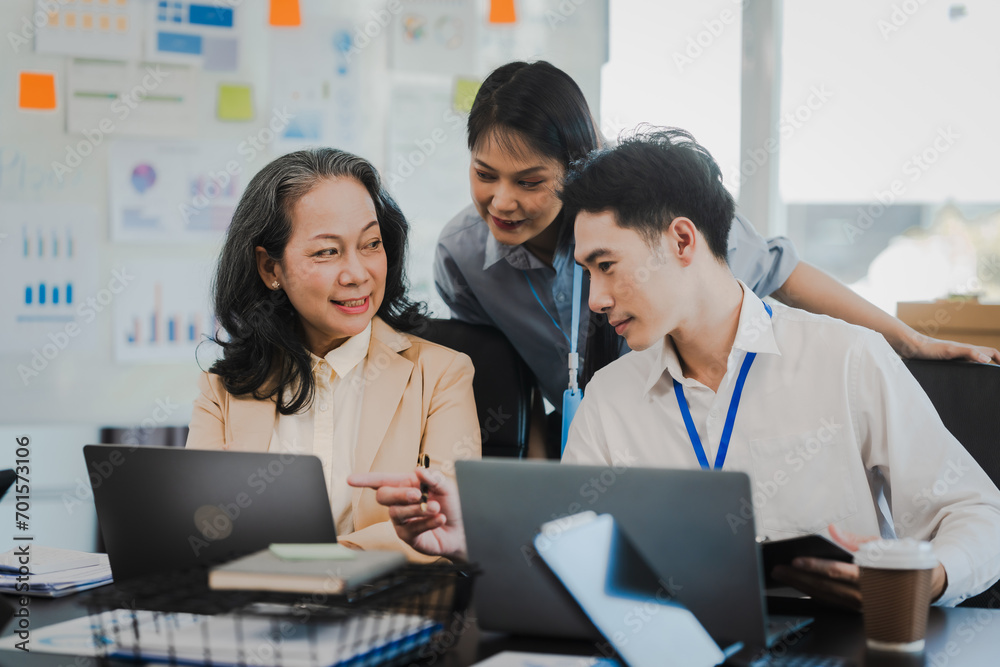 Asian office worker discussing with younger colleagues over laptops in a meeting room, possibly reviewing monthly reports or sharing opinions.