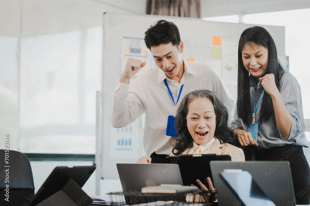 Asian office worker discussing with younger colleagues over laptops in a meeting room, possibly reviewing monthly reports or sharing opinions.