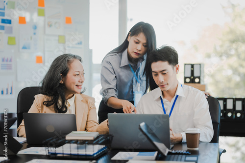 Asian office worker discussing with younger colleagues over laptops in a meeting room  possibly reviewing monthly reports or sharing opinions.