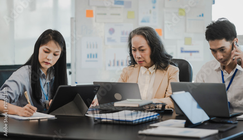 Asian office worker discussing with younger colleagues over laptops in a meeting room, possibly reviewing monthly reports or sharing opinions. © makibestphoto