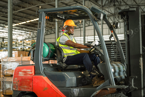 African forklift driver focused on carefully transporting stock from shelves of a large warehouse wearing a helmet and vest looking toward goods