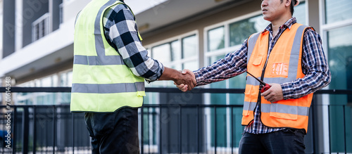 two construction workers, one Asian, shaking hands on a building site, possibly indicating agreement or completion of a task, with a modern building in the background.