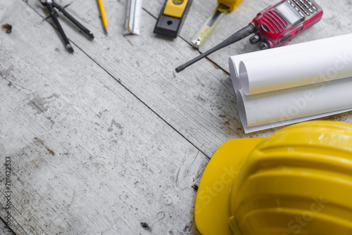 Top view construction tools such as a yellow hard hat, spirit level, measuring tape, folding ruler arrayed against a wooden plank background.