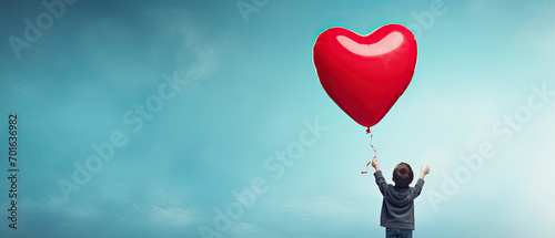 Back view of a kid raising arms with red love valentine heart shaped balloon against sky background