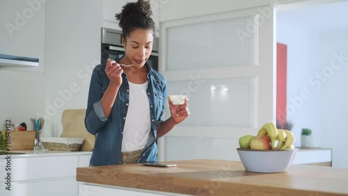 Smiling woman eating yoghurt and checking text messages in kitchen photo