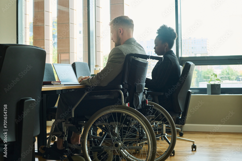 Businessman sitting in wheelchair and using laptop with businesswoman at desk Businessman sitting in wheelchair and using laptop with businesswoman at desk