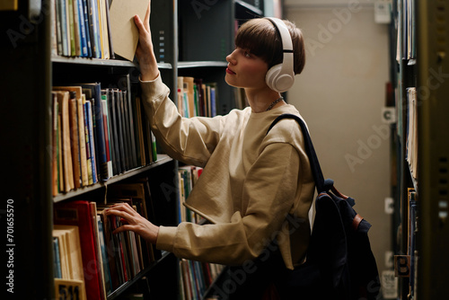 Student wearing wireless headphones and taking book from shelf in library photo