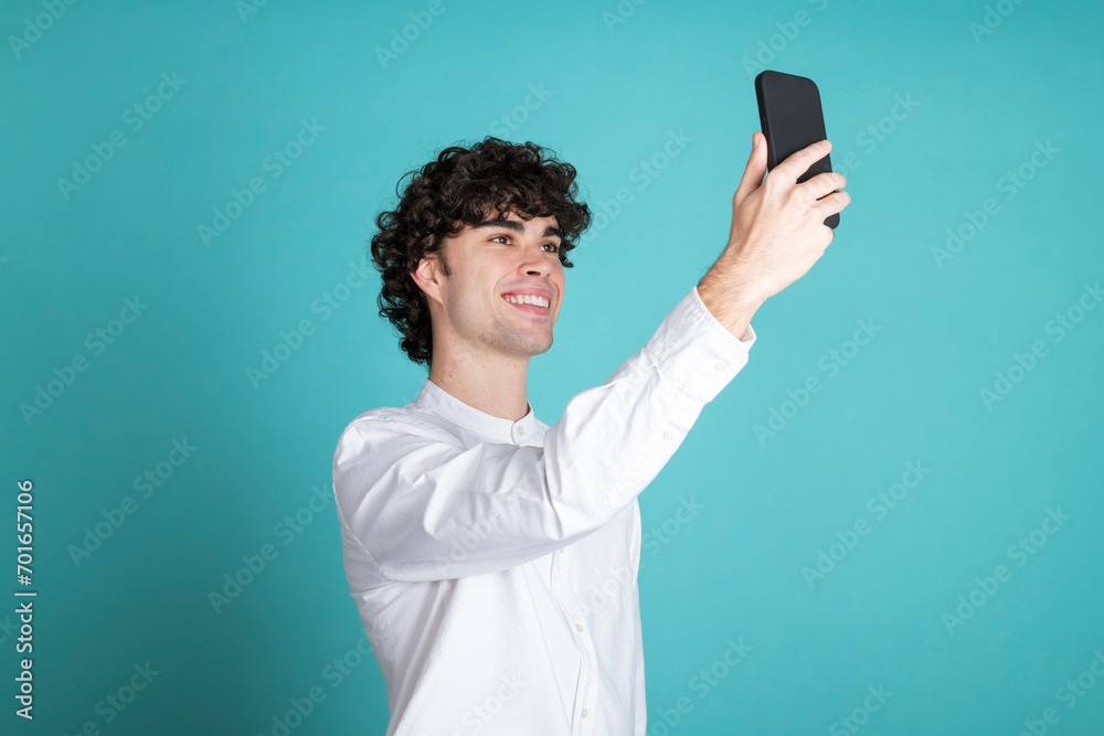 Smiling young man taking selfie through smart phone against cyan background Smiling young man taking selfie through smart phone against cyan background