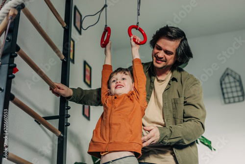 Smiling father teaching son to hang on gymnastic rings at home photo