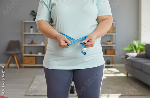 Close up cropped photo of a fat overweight woman wearing sportswear measure the waist with measuring tape standing in the living room at home. Obesity, weight control, diet and slimming concept. photo