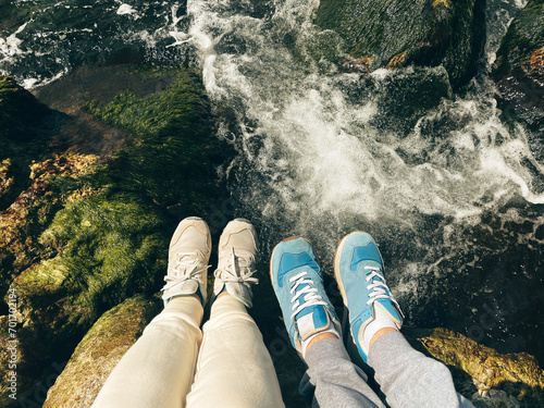 Feet in sneakers on the rocks, selfie over a fast river in the background. Top view of female and male legs over bubbling water. Tourists on a hike.