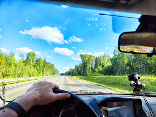 Car salon, steering wheel, hand of woman and view on nature landscape. View from seat of driver on Road, forest, blue sky, white clouds at sunny day. Concept of single trip of female traveller