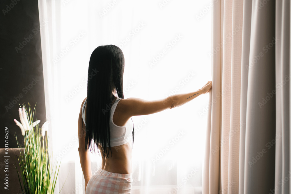 Brunette woman stands at the bedroom window in the morning. She has parted the curtains on the window and is looking at the street Brunette woman stands at the bedroom window in the morning. She has parted the curtains on the window and is looking at the street