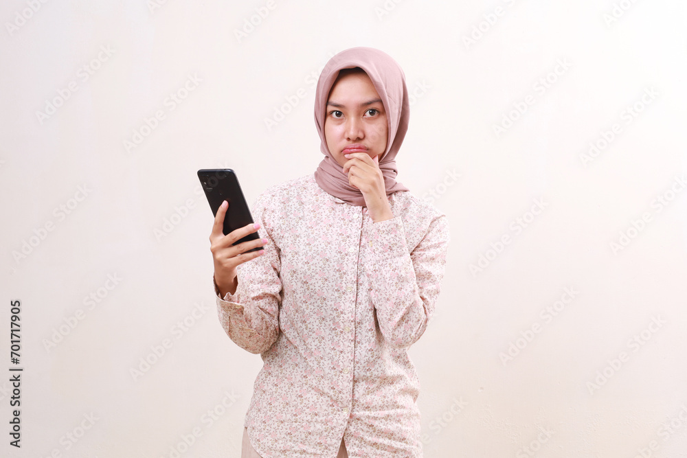 Confused young asian muslim girl standing while holding a cell phone. Isolated on white
