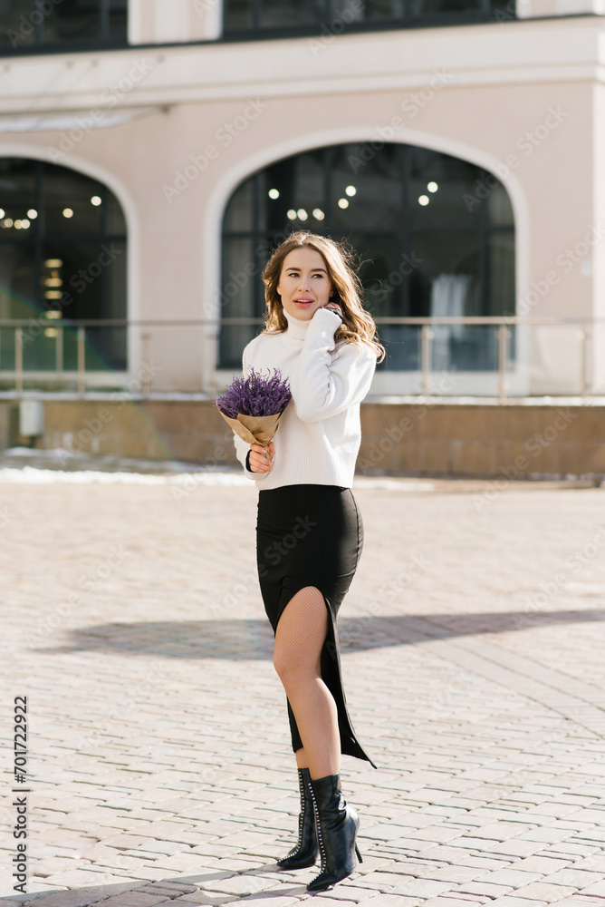 Young Caucasian woman holds a purple bouquet of flowers in her hands and walks along a city street