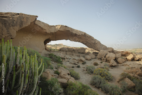 A stunning natural rock arch in a desert landscape, surrounded by arid vegetation and rocky terrain under a clear sky.