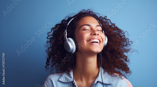 An enticing, joyful woman is posing in a cheerful mood while listening to music on headphones in a checkered shirt and jeans isolated on a blue studio background with an eye aimed upwards.