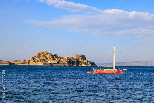 A red sailboat anchors in the blue sea in front of the fortress of Corfu town in the light of the evening sun under a blue sky with light clouds