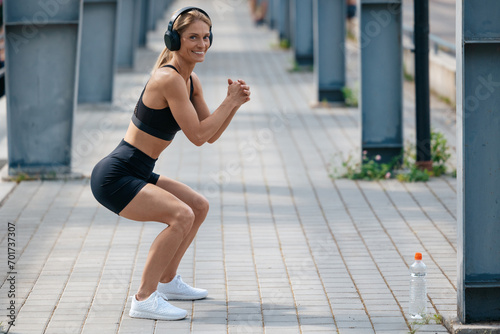 Woman with headphones listening music squatting outdoor doing fitness exercises in summer morning.