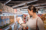A young and beautiful Asian girl is shopping in a supermarket, picking fresh milk from the dairy shelves. She eats healthy, nutritious, and has a balanced diet and lifestyle