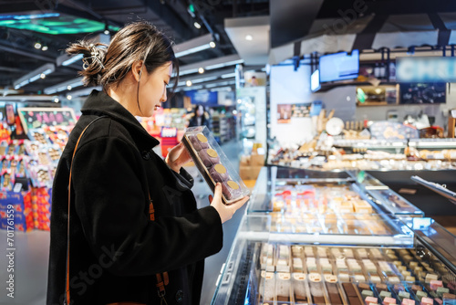 A young Asian girl is shopping in the food section of a supermarket, reading the nutritional labels of fresh baked desserts