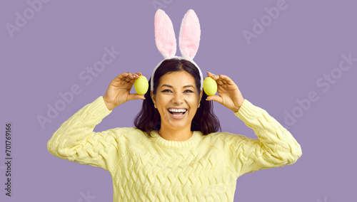 Happy Easter. Cheerful funny woman with bunny ears and with easter eggs on pastel purple background. Happy beautiful woman holding two yellow Easter eggs and sincerely smiling at camera. Banner. photo